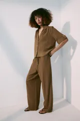 Pantalón de tiro medio color marrón, con cintura elástica y cordón ajustable con abalorios decorativos. Presenta bolsillos laterales y perneras rectas con diseño texturizado.