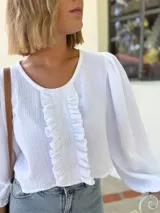 Blusa blanca de algodón con textura arrugada, cuello redondo, mangas largas abullonadas con puños elásticos y cierre frontal con botones y volantes.