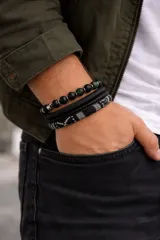 Set de tres pulseras para hombre. Una pulsera de cuentas negras mate con detalles de piedras verdes y cuentas metálicas plateadas. Dos pulseras de cuero sintético negro con diferentes texturas. Una pulsera de tela trenzada con diseño geométrico en blanco y negro.