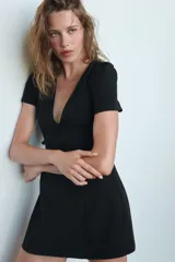Vestido corto de color negro con escote en pico profundo, mangas cortas abullonadas y detalle de lazo frontal. Presenta un diseño entallado con cierre de cremallera oculto en la espalda.