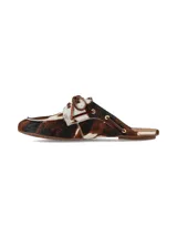 Zueco de cuero con pelo con estampado animal print tipo vaca en tonos marrones, blancos y negros. Presenta un diseño destalonado con cordones decorativos en la parte superior y ojales metálicos.