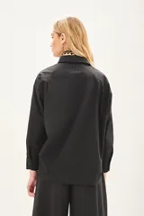 Camisa de popelina de algodón en color negro, con corte amplio, cuello clásico y cierre frontal con botones. Presenta un diseño versátil con botones en los laterales que permiten un ajuste cruzado.