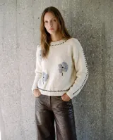 Buzo tejido de lana merino, color crudo con flores bordadas grises y pespuntes negros a contraste en cuello, hombros y ruedo, de manga larga y calce relajado.