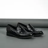 Mocasín de cuero vacuno negro con textura grabada tipo reptil. Presenta un diseño clásico con antifaz en el empeine, suela de goma en tono natural y taco bajo de madera.