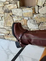Bota de caña alta estilo biker, confeccionada en cuero color chocolate. Presenta un cierre metálico lateral que recorre toda la caña, estribo y cadena decorativa desmontables, y taco cuadrado con plataforma.