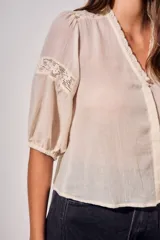 Blusa de color arena con cuello en V, mangas tres cuartos abullonadas y detalles de encaje bordado en hombros y mangas. Presenta un diseño ligero y delicado con cierre frontal.