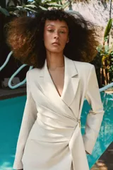 Blazer de corte estructurado en color beige, con cuello solapa y manga larga. Presenta un diseño cruzado con cierre mediante lazada del mismo tejido y un distintivo aplique metálico dorado en el frente.