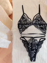 Conjunto de lencería compuesto por un bralette triangular y una tanga, confeccionados en encaje negro con detalles florales en color hueso. El bralette presenta breteles ajustables y diseño con tiras decorativas, mientras que la tanga cuenta con doble tira lateral regulable.