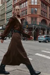 Vestido midi de color marrón con diseño combinado, escote en pico con volantes, mangas largas con puños elásticos y falda evasé. Incluye un cinturón negro con hebilla dorada en la cintura y cierre lateral con cremallera oculta.