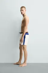 Short de baño blanco marfil con cintura marrón y cordón ajustable del mismo color. Presenta franjas laterales en tono azul marino con finos bordes rojizos y bolsillos laterales.