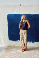 Pantalón ancho de tiro alto color beige, con pliegues frontales y botones visibles en la cintura. El ruedo del pantalón tiene un detalle de terminación en cuero color caramelo.