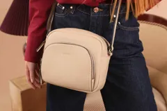 Bandolera de diseño compacto en color beige, confeccionada en material símil cuero con textura granulada. Cuenta con un bolsillo frontal con cierre y correa larga regulable.