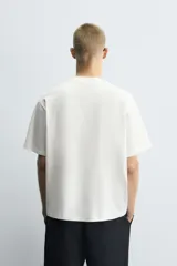Camiseta boxy fit de color blanco con cuello redondo y mangas cortas. Presenta un parche rectangular en el centro del pecho con texto impreso y costuras decorativas a contraste en los laterales.