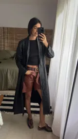 Tapado largo estilo trench de cuero ovino negro, con silueta recta, solapa amplia y cierre frontal cruzado ajustable con lazo en la cintura. Cuenta con bolsillos laterales funcionales y largo midi por debajo de la rodilla.
