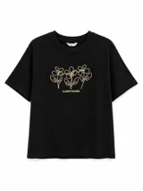 Remera de algodón color negro, con cuello redondo y mangas cortas. Presenta un diseño lineal bordado en color beige que representa tres flores y el texto 'FLOWER PARADISE' en la parte central del pecho.