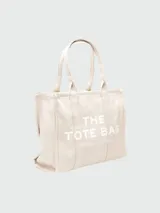 Bolso tipo tote de Marc Jacobs, confeccionado en lona resistente de color negro. Presenta el nombre del modelo estampado en blanco en la parte frontal y el logo de la marca debajo. Cuenta con doble asa superior para llevar al hombro o en la mano.