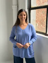 Sweater de punto color azul lavanda, con escote en V, mangas largas con puños acanalados y dos bolsillos frontales tipo parche. Presenta una costura central vertical visible.