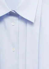 Camisa de popelín de algodón en color celeste claro, con cuello camisero, cierre frontal de botones y detalle de lazada tipo corbata integrada al cuello. Presenta un bolsillo de parche en el pecho, corte recto y mangas largas con puños abotonados.