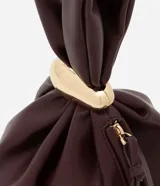 Bolso tipo bucket pequeño color marrón oscuro, confeccionado en material sintético con poliéster certificado. Tiene correa de mano y detalle de metal dorado.