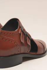 Zapato tipo monk strap con aberturas laterales, confeccionado en cuero vacuno metalizado color bronce. Presenta detalles de perforaciones estilo brogue en la puntera y el talón, y doble hebilla metálica en el empeine.