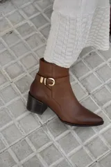 Bota corta de cuero color marrón, con taco cuadrado de madera y punta fina. Presenta una tira decorativa con hebilla metálica dorada alrededor del tobillo.