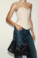 Bolso tipo saca color negro, de diseño flexible y acabado con efecto gastado. Cuenta con un asa ajustable para el hombro, detalles de hebillas y herrajes metálicos laterales, además de un cierre superior de cremallera metálica.
