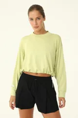 Buzo crop color verde claro, con cuello redondo y mangas largas holgadas. Presenta frunce en el ruedo mediante cordón elástico y tanca.