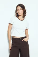 Remera cropped blanca de manga corta, con cuello redondo y terminaciones en contraste de color marrón en cuello y mangas. Inspirada en los clásicos de los años 80.