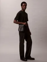 Morral Calvin Klein modelo Cargo Reporter, color negro, confeccionado en poliuretano con diseño estructurado, solapa frontal y cierre de hebilla. Cuenta con correa de hombro ajustable y detalles de la marca grabados.
