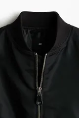 Campera bomber de nylon color negro, con cuello, puños y bajo acanalados. Presenta cierre frontal, bolsillos laterales con botón a presión y un bolsillo con cierre en la manga.