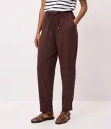 Pantalón carrot marrón de tiro alto, confeccionado en mezcla de viscosa y poliéster, con cintura elástica fruncida, cordón de ajuste tipo lazo con punteras metálicas, bolsillos y recortes estructurados en las piernas.