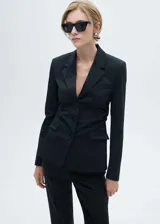 Blazer entallado de color negro con cuello de solapa, cierre frontal de tres botones y bolsillos delanteros con solapa. Presenta un diseño estructurado con detalles fruncidos en los laterales y forro interior.