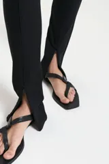 Pantalón negro de viscosa con calce al cuerpo, tiro medio y largo a los talones. Presenta cintura con elástico interno en la espalda, detalle de tira cruzada con medialunas metálicas, sustracción en el frente de la cintura, recortes verticales en el delantero, espalda con bolsillo falso ojal y pinzas en la cintura, y tajos en el ruedo con cierre invisible.