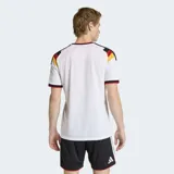 Camiseta de fútbol Adidas de la primera equipación de Alemania, modelo 26. Diseño principal blanco con un patrón en V en el pecho que incorpora los colores de la bandera alemana (negro, rojo y amarillo/dorado). Posee cuello en V con ribete rojo y detalles en las mangas. El escudo de la federación alemana con cuatro estrellas está bordado en el pecho, junto al logo de Adidas.