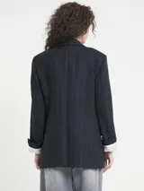 Blazer de corte recto, color negro o azul muy oscuro, con finas rayas verticales en tono claro (posiblemente marrón o beige). Presenta solapas de muesca, cierre frontal de dos botones y bolsillos de parche en la parte inferior. Se lleva con una camisa blanca a rayas finas asomando por las mangas.