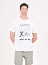 Remera blanca de manga corta con cuello redondo y estampado con la frase "New York City Tennis 1989" y una ilustración de dos jugadores de tenis.