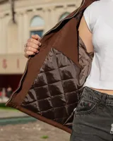 Chaqueta de estilo utilitario color marrón, con cierre frontal completo, cuello camisero, dos bolsillos frontales tipo parche y un bolsillo pequeño con cierre en el pecho. Presenta un bordado decorativo en el pecho y puños con botones.