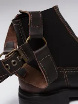 Bota de cuero color marrón oscuro con punta cuadrada, diseño tipo chelsea con paneles elásticos laterales y estribo decorativo desmontable con hebilla metálica.