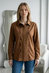 Sobrecamisa de gamuza color camel, con cuello clásico, cierre frontal con botones metálicos, detalles de tachas a lo largo de la botonadura y bordados florales verticales. Presenta mangas largas con puños abotonados y tachas decorativas.