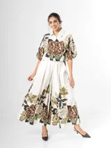 Conjunto de dos piezas confeccionado en viscosa con estampado floral en tonos marrones y verdes sobre base blanca. Incluye una blusa tipo camisa corta con cuello solapa, cierre frontal de botones y mangas abullonadas con diseño globo, y un pantalón tipo culotte de tiro alto, amplio y fluido con pinzas en la cintura.