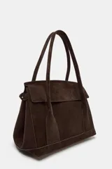 Bolso tipo citybag confeccionado en serraje color marrón oscuro. Presenta una solapa frontal con cierre magnético, asas de hombro integradas y detalles de hebillas metálicas en los laterales para ajuste. Incluye bolsillo interior con cremallera.