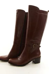 Bota de caña alta confeccionada en cuero legítimo color marrón, con diseño clásico, costuras visibles y una delicada tira con hebilla lateral. Cuenta con cierre lateral interno y taco medio ancho.