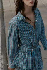 Vestido corto de manga larga con cuello solapa y cierre frontal de botones a presión. Presenta un diseño a cuadros en tonos azul y marrón, cintura ajustable con cinturón de hebilla y falda con volantes en el bajo.
