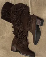 Bota bucanera de caña alta confeccionada en cuero vacuno color marrón, con diseño estilo boho que presenta flecos largos a lo largo de la caña y taco cuadrado de madera.