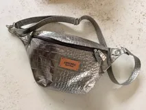 Riñonera de cuero sintético con textura croco en color plateado, cierre frontal marrón y correa ajustable.