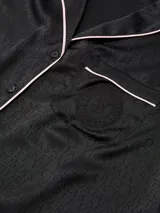 Pijama largo de satín negro, compuesto por una camisa de manga larga con botones, cuello con muesca, bolsillo en el pecho y ribete blanco, y un pantalón largo a juego con cintura elástica.