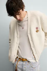 Cárdigan de punto color beige con cuello en pico, manga larga y cierre frontal de botones. Presenta un pequeño bordado a contraste en el pecho y acabados en rib en puños y bajo.