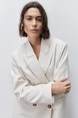 Blazer de silueta amplia y largo por debajo de la cadera, color marfil, confeccionado en tela suave y liviana. Presenta cuello con solapas, hombreras, manga larga y cierre cruzado con botones carey.