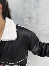 Campera tipo gamulán de color negro con interior y detalles de corderito en color blanco. Presenta cierre frontal, cuello clásico y dos bolsillos frontales.