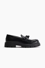 Mocasines negros de suela gruesa con costura frontal y detalle de tiras decorativas cruzadas con nudos.